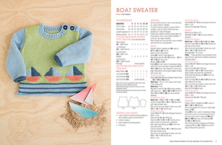 Hand-knitted baby sweater with sailboat design on a light wood background, next to small paper boats and sand. Instructions labeled "Boat Sweater."