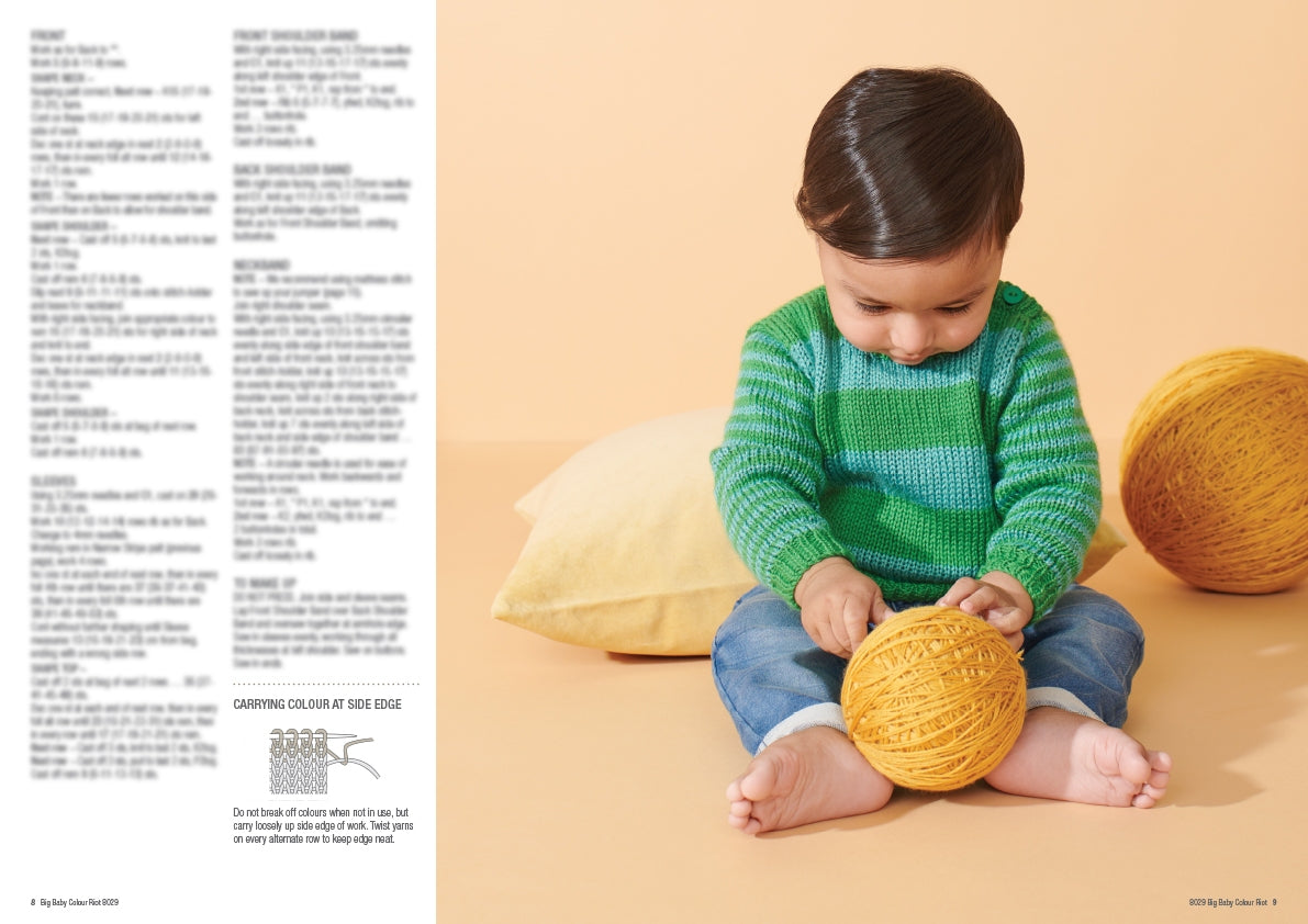 A baby in a green striped sweater and jeans sits barefoot on a peach background, playing with a large yellow yarn ball, next to a yellow cushion.