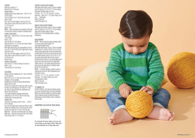 A baby in a green striped sweater and jeans sits barefoot on a peach background, playing with a large yellow yarn ball, next to a yellow cushion.