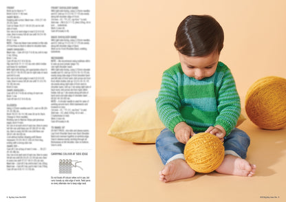 A baby in a green striped sweater and jeans sits barefoot on a peach background, playing with a large yellow yarn ball, next to a yellow cushion.