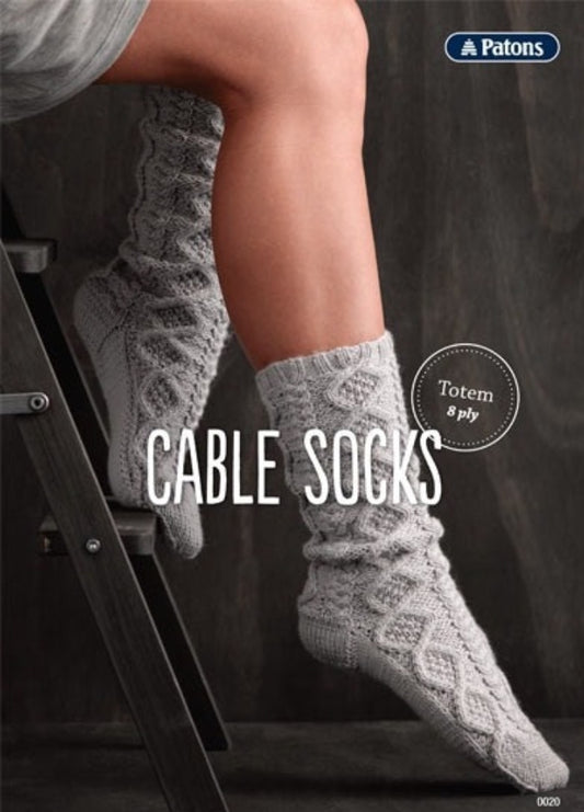 A person sits on a ladder wearing gray cable-knit socks. The background is dark gray. Text reads "Cable Socks" and "Totem 8 ply" with a Patons logo.