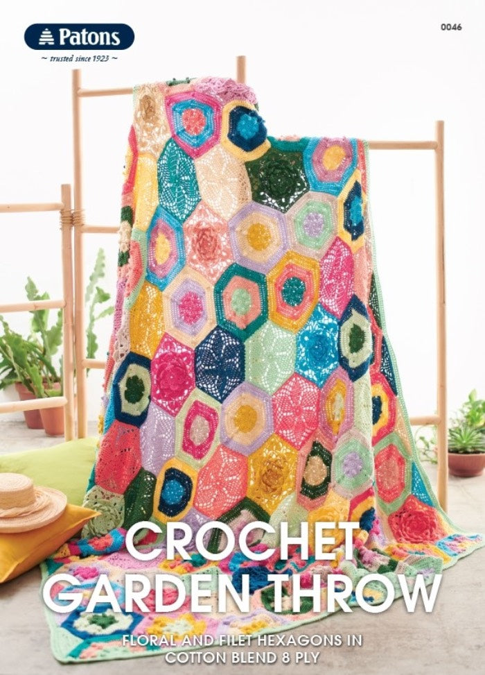 Colourful crochet blanket with hexagonal patterns draped over a wooden stand, set against a minimalist background with potted plants and a straw hat. Cozy and vibrant.