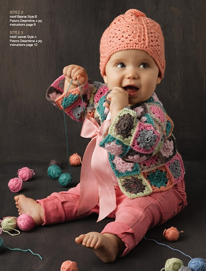A baby wearing a colorful crocheted sweater and pink hat sits on the floor, surrounded by various colored yarn balls, holding a string and smiling.
