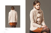 A woman in a cozy cream knit sweater with pom-pom accents sits on a stool. She pairs it with a brown skirt. The mood is warm and stylish.