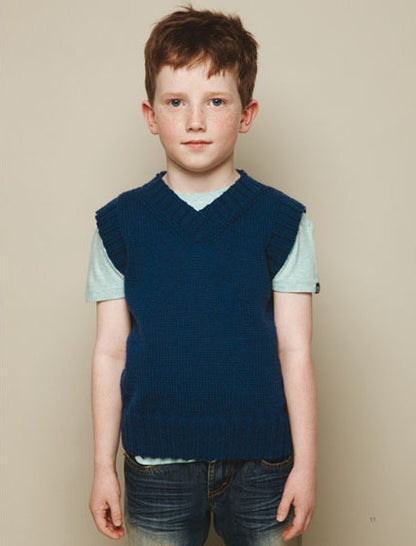 Young boy with short brown hair, wearing a light blue shirt and dark blue knitted vest, stands against a plain beige background, looking calm and composed.