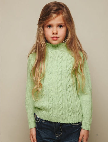 Young girl with long, blonde hair wearing a light green cable-knit sweater and dark jeans stands against a neutral background, gazing seriously.