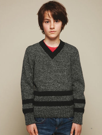 Young person with short brown hair wearing a gray V-neck sweater with black stripes and blue jeans, standing against a plain beige background, appearing calm.