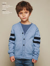 A young child with brown hair wearing a light blue cardigan with dark blue stripes on the sleeves. The child stands against a plain background.