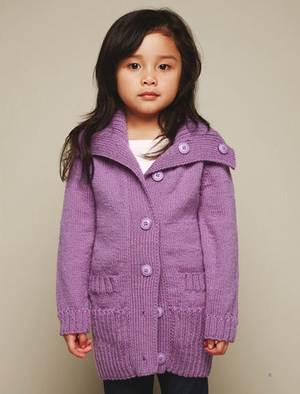 A young girl with long dark hair wears a lavender knitted cardigan with large buttons. She stands against a plain beige background, looking calm and composed.