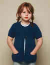 Young child with brown hair in braids, wearing a blue knitted cardigan with short sleeves and pockets, stands against a neutral background, looking calm.