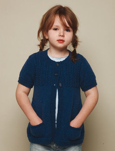 Young child with brown hair in braids, wearing a blue knitted cardigan with short sleeves and pockets, stands against a neutral background, looking calm.