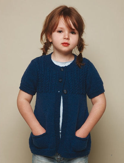 Young child with brown hair in braids, wearing a blue knitted cardigan with short sleeves and pockets, stands against a neutral background, looking calm.
