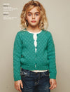Young girl with curly blonde hair wearing a teal knitted cardigan over a white top and denim jeans, standing against a beige background, looks calm.