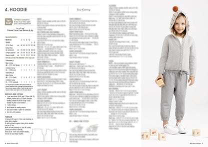 Child in gray knit hoodie with pom-pom and striped hood, holding a block. Standing on white surface with scattered wooden alphabet blocks. Cozy and playful.