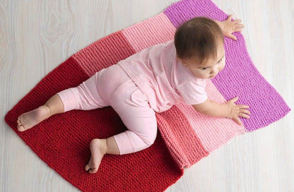 A baby in pink clothes lies on a colorful knitted blanket with shades of red, pink, and purple. The light wooden floor adds a soft, cozy atmosphere.