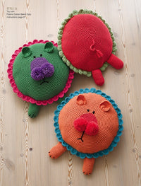Three colorful crochet lion cushions on a wooden floor. One is green with a purple nose, another red with green edging, and an orange one with blue edging. Each has a friendly expression.