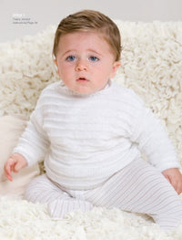 A baby with light brown hair sits on a cream, textured blanket, wearing a white knitted sweater and striped pants, looking upward with a curious expression.