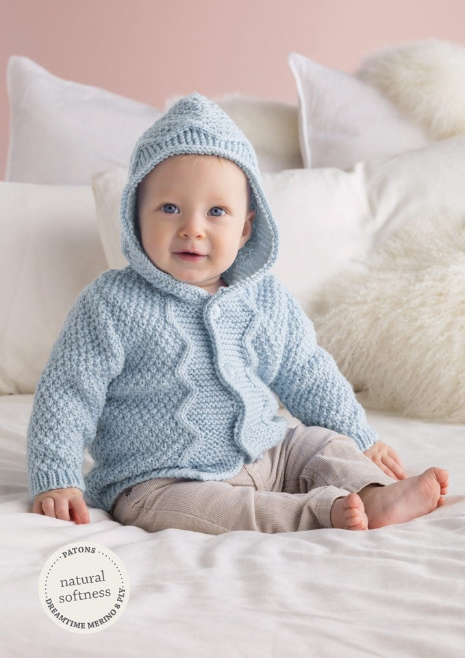 A baby in a cozy blue knit hoodie sits on a bed with white pillows and a pink wall. The scene conveys warmth and softness. A "natural softness" label is visible.