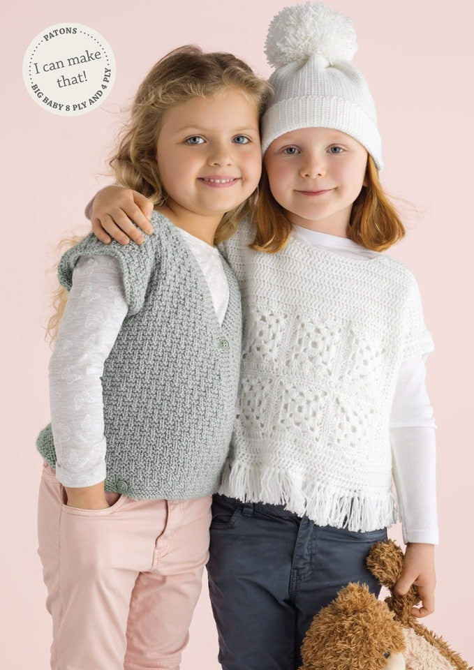 Two young children smile while posing together against a soft pink background. One wears a knitted gray vest, the other in a white sweater and pom-pom hat.