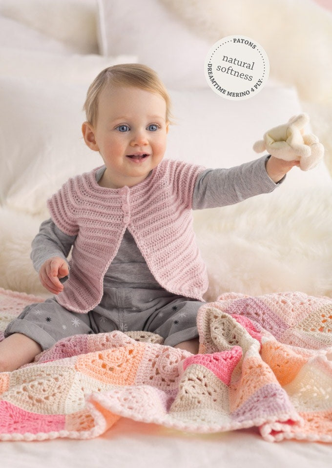A cheerful baby in a pink knit cardigan sits on a colorful crocheted blanket, holding a toy. The setting is cozy and soft with a warm tone.