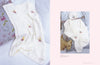 A soft white knitted baby blanket with floral embroidery rests on a floral pillow, paired with a matching cardigan and hat on a bed, exuding warmth and coziness.