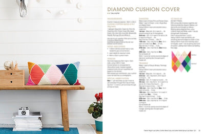 A cozy interior with a wooden bench holding a colorful diamond-patterned crochet cushion. A white wall background, books, camera, and red flowers add charm.