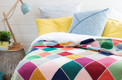 Brightly colored geometric-patterned blanket on a bed with yellow and blue pillows. A wooden lamp and small potted plant sit on a rustic bedside table.