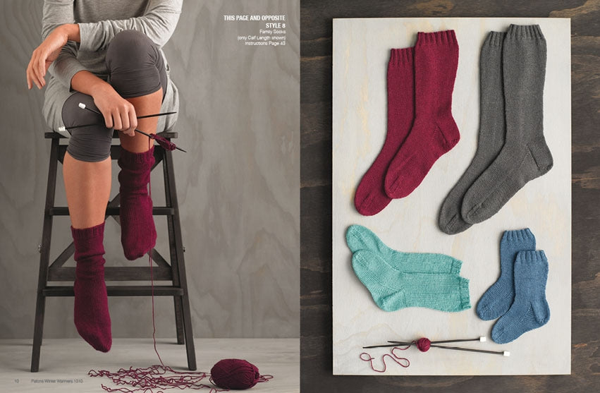 A person sitting on a ladder is knitting burgundy socks, with yarn and needles nearby. On the right, burgundy, gray, teal, and blue socks are displayed on a board.