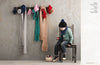 A child in winter clothes sits on a chair, holding a red toy. Colorful scarves and hats hang on the wall. The scene feels cozy and playful.