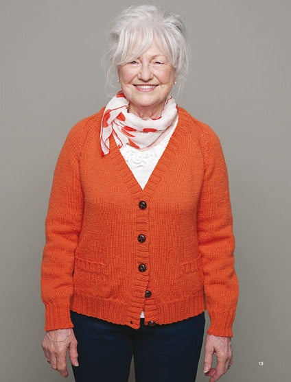 Elderly woman smiling, gray hair styled up, wearing an orange cardigan with white button-up blouse and patterned scarf, set against a plain gray background.