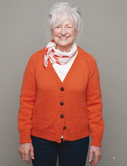 Elderly woman smiling, gray hair styled up, wearing an orange cardigan with white button-up blouse and patterned scarf, set against a plain gray background.