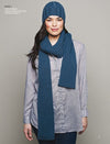 A woman in a blue knit hat and matching scarf over a light gray button-up shirt stands against a plain background, conveying a cozy, casual vibe.