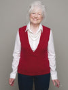 Elderly woman with white hair smiles warmly, wearing a red sweater vest over a white shirt and blue pants, set against a plain gray background.