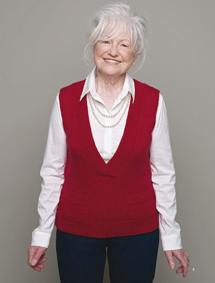 Elderly woman with white hair smiles warmly, wearing a red sweater vest over a white shirt and blue pants, set against a plain gray background.