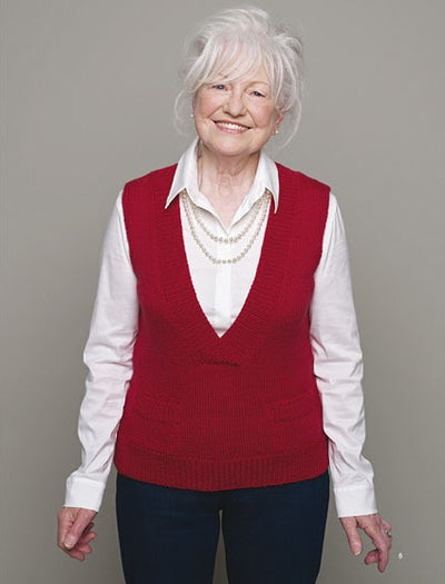Elderly woman with white hair smiles warmly, wearing a red sweater vest over a white shirt and blue pants, set against a plain gray background.