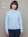 A woman in a light blue sweater and jeans stands against a gray background. She smiles softly, exuding a warm and cozy mood.