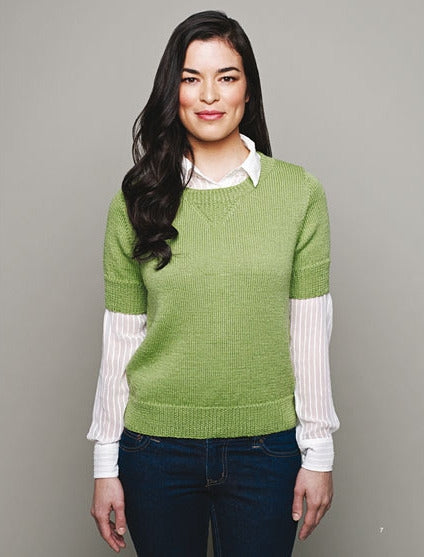 Smiling woman in green sweater over a white collared shirt and blue jeans against a gray background. Long dark hair, relaxed and confident demeanor.