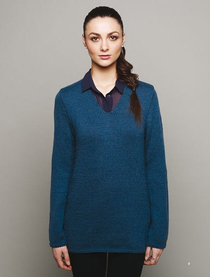 A person with a calm expression stands against a gray background, wearing a teal sweater over a dark collared shirt. The mood is neutral and composed.
