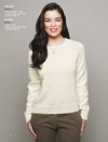 Smiling woman wearing an ivory knit sweater and olive pants, standing against a gray background. Her long, dark hair is styled in waves.