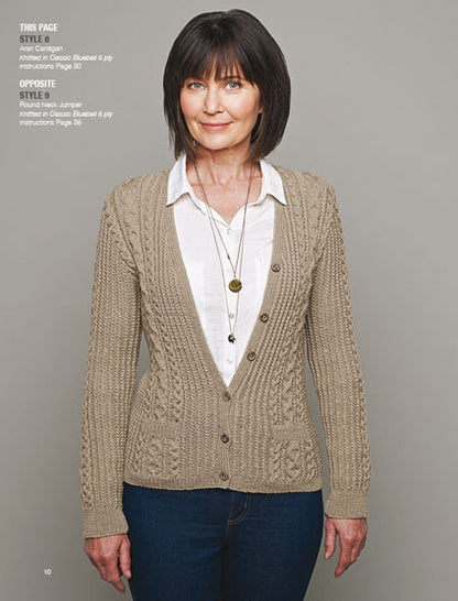 Woman wearing a beige knit cardigan over a white shirt, with long necklaces, standing against a gray background. She has a calm and confident expression.