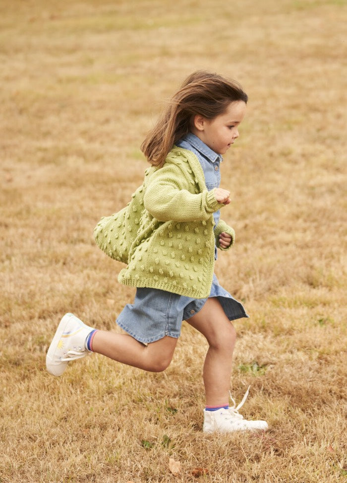 A young child wearing a green knit sweater, denim dress, and white sneakers joyfully runs across a dry grassy field, exuding energy and playfulness.