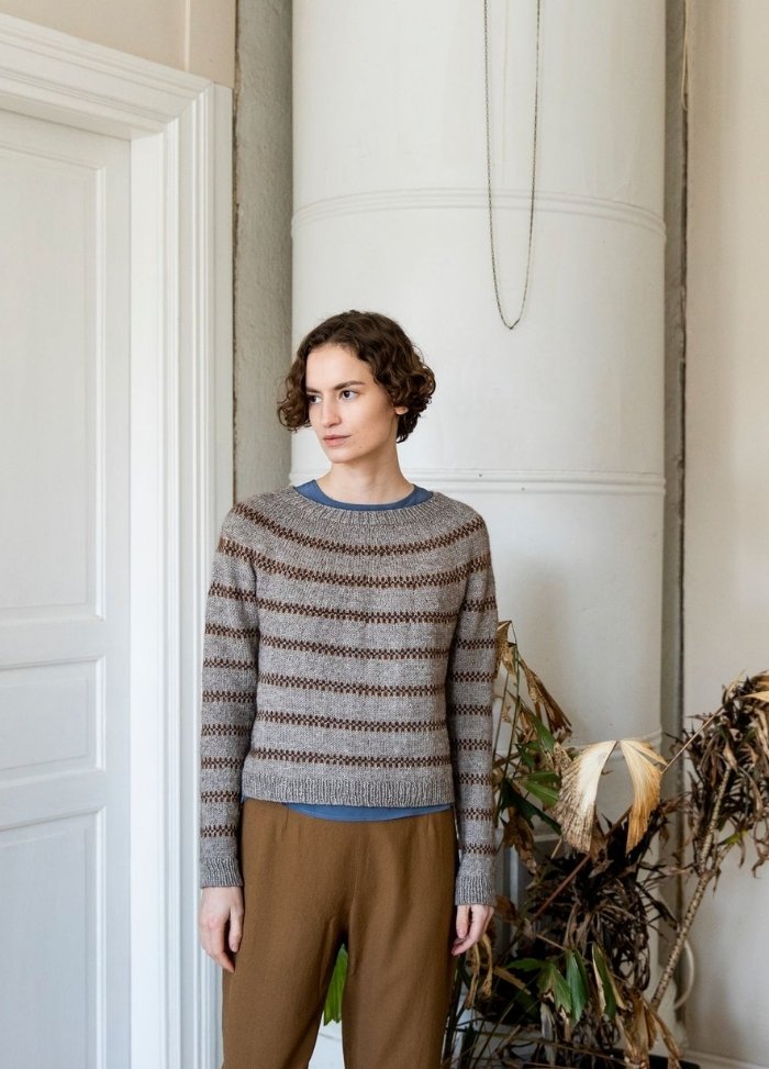 Alen Sweater: striped knit sweater in brown and beige tones, styled with brown pants and a blue shirt, photographed indoors.