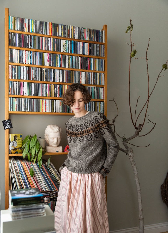 Balaio Sweater: gray knit sweater with patterned yoke, styled with a long skirt, photographed in front of a shelf of CDs and vinyl records.