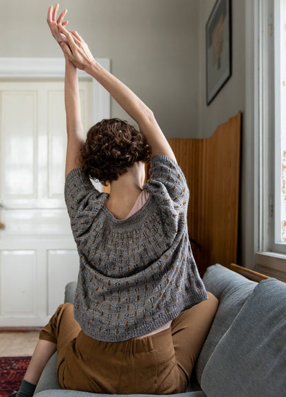 Brocado Sweater: gray sweater with intricate patterns, worn with brown pants, photographed indoors with arms raised on a couch.