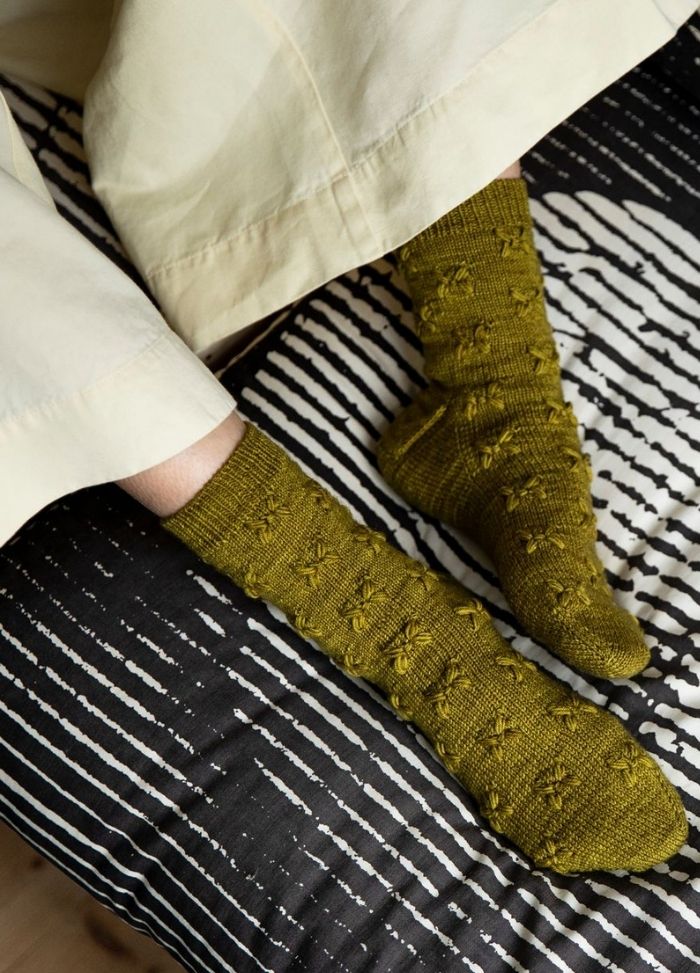 Caliandra Socks: green textured knit socks worn with rolled-up pants, photographed on a striped bedspread.