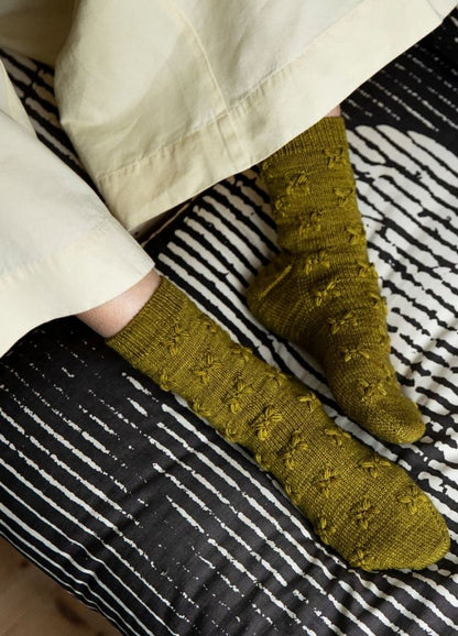 Caliandra Socks: green textured knit socks worn with rolled-up pants, photographed on a striped bedspread.