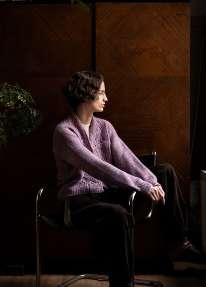 Cestaria Cardigan: purple knit cardigan worn with dark pants, photographed seated against a wooden backdrop with greenery.