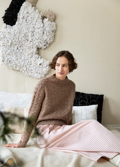 Diamantine Sweater: brown textured knit sweater paired with a pleated striped skirt, photographed seated with abstract textile art in the background.