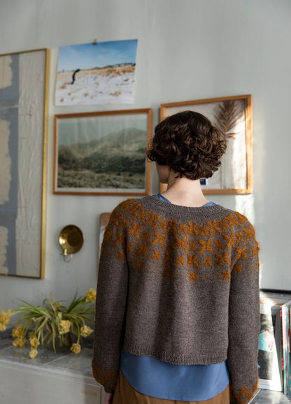 Florada Sweater: gray knit sweater with orange floral yoke, photographed from behind in a room with framed art and yellow flowers.