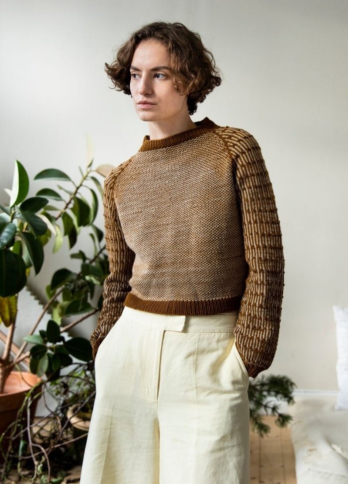 Lina Sweater: brown knit sweater with textured sleeves, worn with beige pants, photographed against a light wall with green plants.